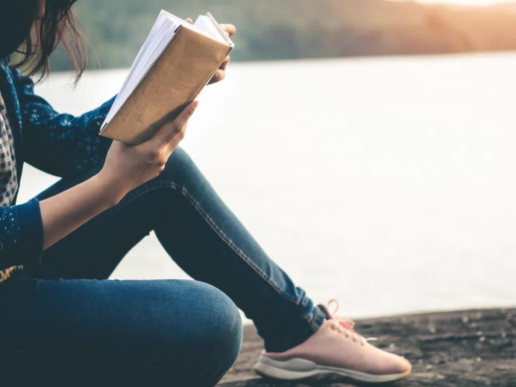 Teen reading book alongside a lake at sunset.