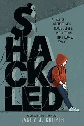 Cover of Shackled by Candy J. Cooper. The letters spelling "Shackled" stacked like blocks. The "S" a dollar sign, a metal linked chain winding through the letters a young person leaning against the letter "D," the chain ending in cuffs on the person's hands.