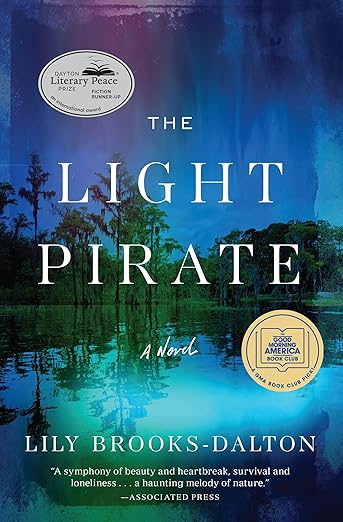 Book cover of “The Light Pirate” by Lily Brooks-Dalton. The artwork shows a glowing blue and green waterscape with reflections of trees along the shoreline. Two award badges appear near the top left and bottom right.