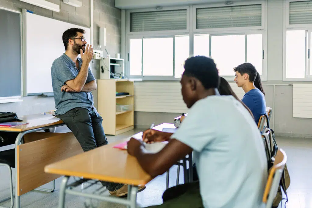 High school students participate in a class discussion while a teacher sits on a desk and gestures, leading a conversation about short stories for students, especially those with unique structures and perspectives.