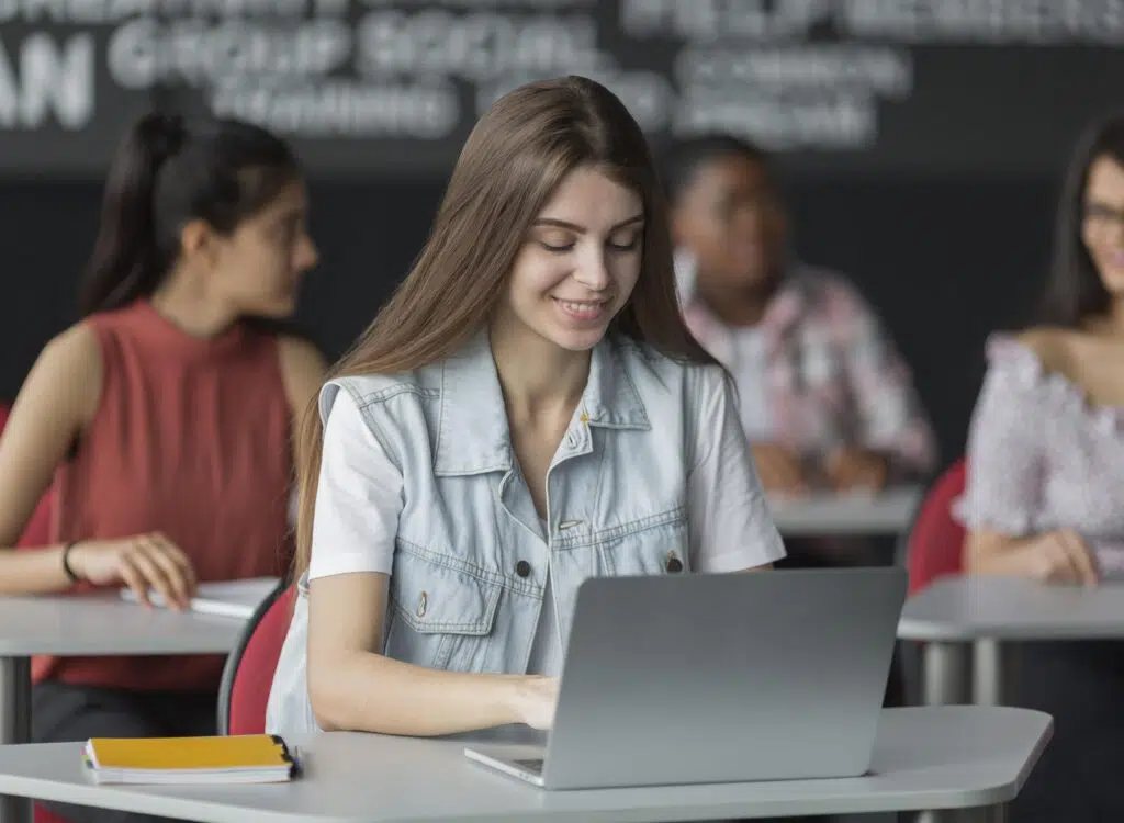 A student works on a laptop during class while other students sit behind her, focused on a writing lesson about business letter format and professional communication skills.
